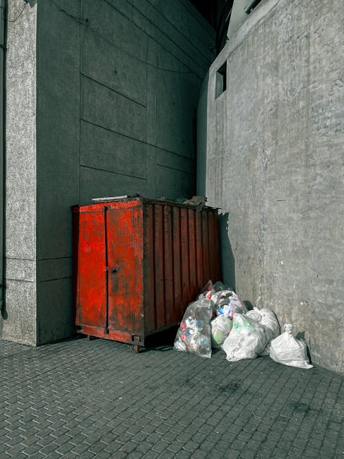 A red metal skip with a weathered and slightly rusted surface is positioned against a modern building's exterior wall, which features dark green panels on one side and light grey concrete on the other