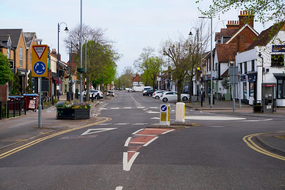 A quiet street scene featuring a pedestrian crossing with white dashed lines and orange textured pavement markings in the foreground. On the left side, a red and yellow give way traffic sign is mounte